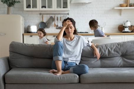 Young tired single mother suffers from headache closed eyes touch forehead sitting on couch while her daughter and son running around her and shouting